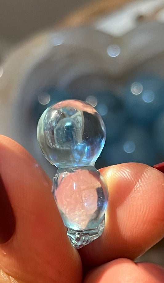 May include: A clear blue glass figurine shaped like a person with a large round head and a smaller round body.